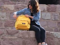 A woman sits on a historic stone wall in Ankara, checking her backpack on a sunny day.